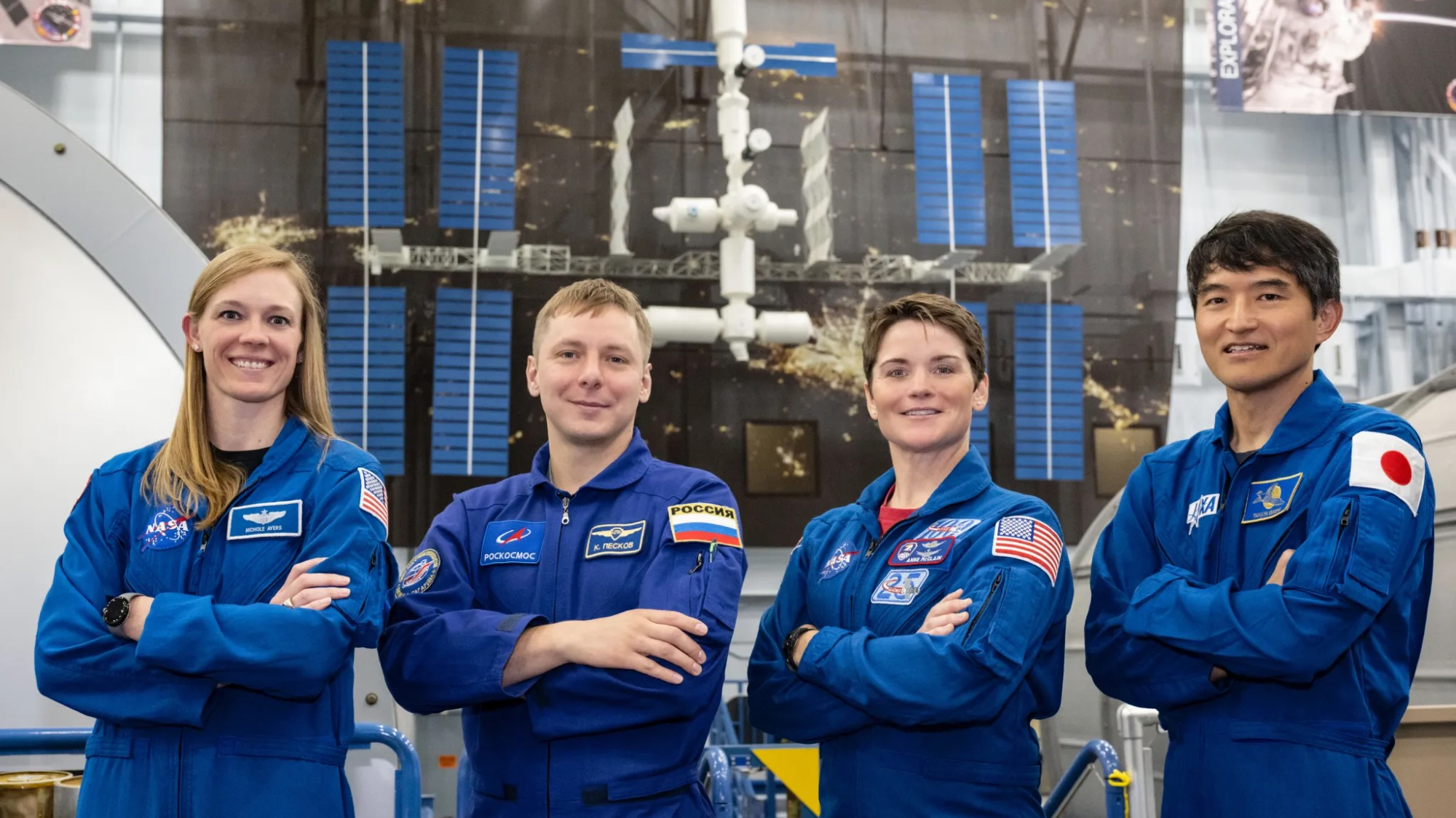 Vier Astronauten in blauen Overalls mit einem Raumstationsmodell hinter sich.