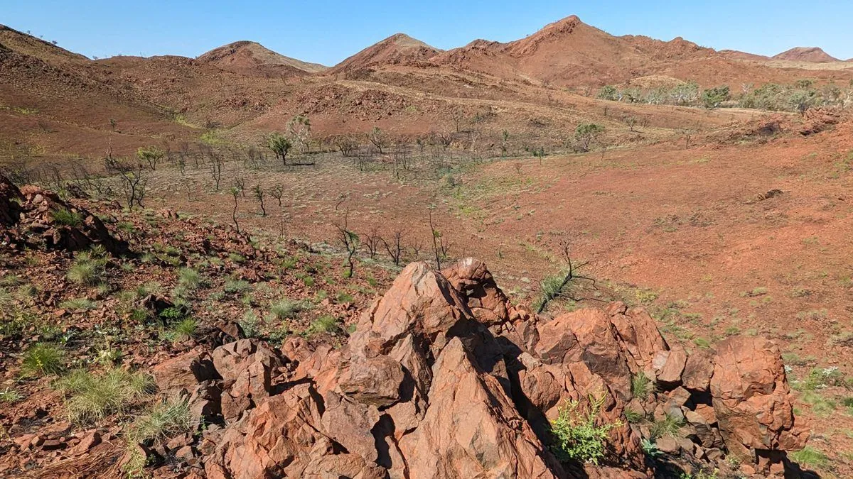 Eine Landschaft mit rötlichen Felsen. Im Hintergrund sind Berge zu sehen, und die Vegetation ist spärlich.