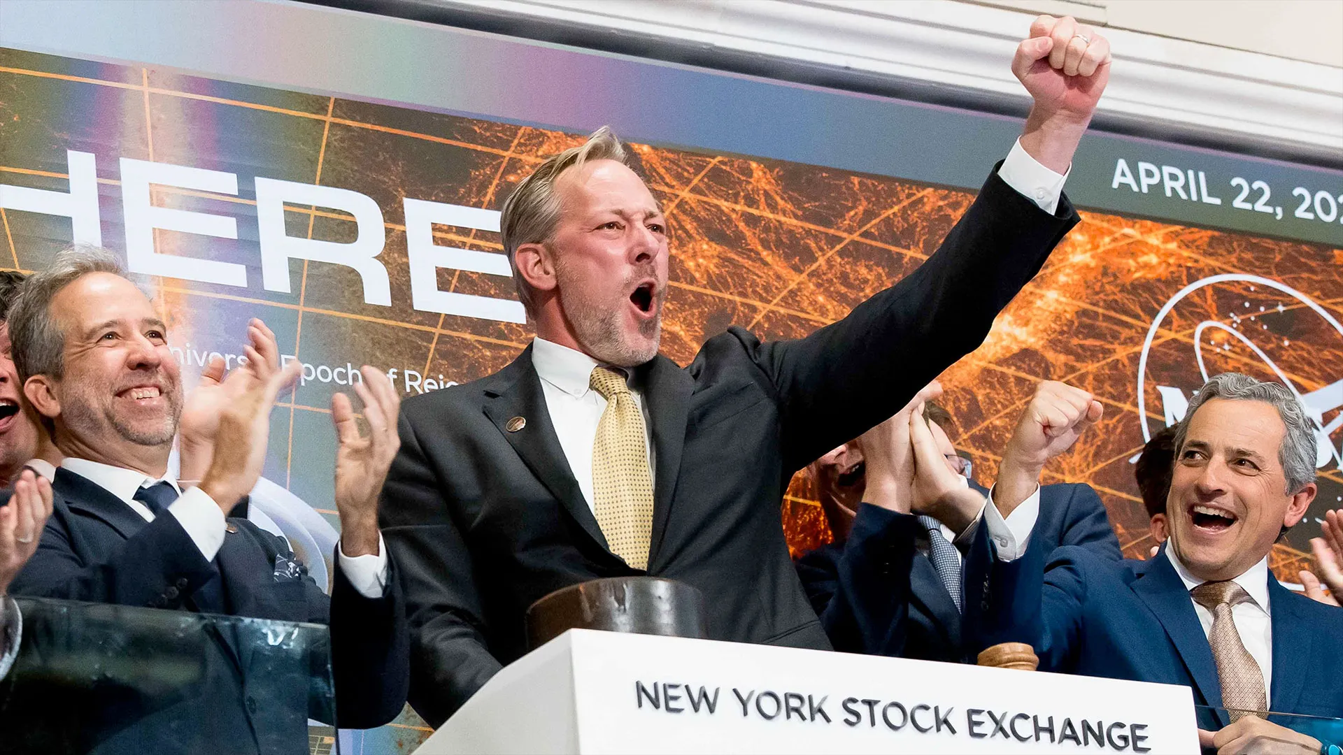 Ein Mann mit schmutzig-blondem Haar und hellem Bart, der einen schwarzen Anzug, ein weißes Hemd und eine hellbraune Krawatte trägt, reckt die rechte Faust in die Luft, als er die Schlussglocke der New Yorker Börse läutet. Andere Männer in Anzügen applaudieren um ihn herum.